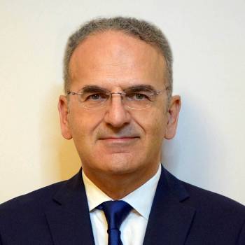 Gordon Crozier DO vilkintis juodą smokingą ir varlytę, stovi priešais banerį su logotipais, įskaitant Oscars ir HeartDaHeart Foundation, oficiali renginio aplinka, neutralus ir profesionalus įspūdis, matomas tekstas: Oscars HeartDaHeart Foundation UnmaskHeartDisease